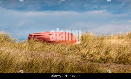 Hohes Gras am Strand, orangefarbenes Boot im Gras mit dem Boden nach oben. Blauer Himmel mit schönen Wolken. Stockfoto