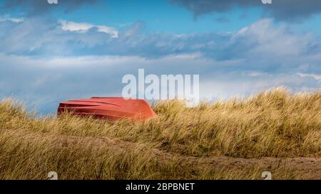 Hohes Gras am Strand, orangefarbenes Boot im Gras mit dem Boden nach oben. Blauer Himmel mit schönen Wolken. Stockfoto