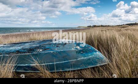 Hohes Gras am Strand blaues Boot liegt im Gras mit dem Boden nach oben. Das Meer im Hintergrund. Blauer Himmel mit großem Schieber. Stockfoto