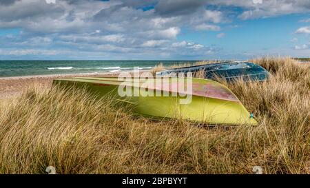 Hohes Gras am Strand, grüne und blaue Boote liegen im Gras mit dem Boden nach oben. Das Meer im Hintergrund. Blauer Himmel mit schönen Wolken. Stockfoto