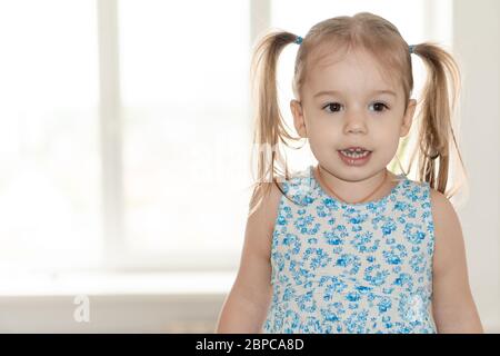 Kindheit, Menschen, Emotionen Konzept - mittel Plan kleines Mädchen mit zwei Pferdeschwänzen und einem blauen Kleid lächelt und zeigt Emotionen auf dem Hintergrund der Stockfoto