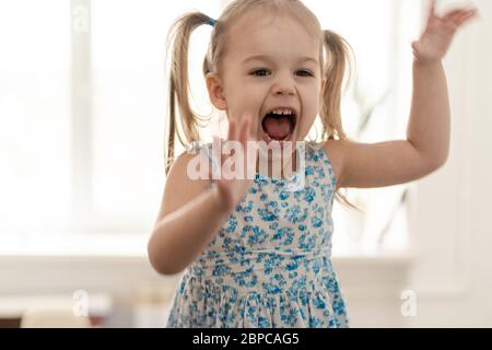 Kindheit, Menschen, Emotionen Konzept - mittelgroßer Plan kleines Mädchen mit zwei Pferdeschwänzen und einem blauen Kleid springt die Hände hoch und schreit am Fenster. Stockfoto