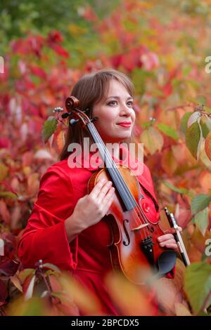 Junge schöne Frau in rotem Mantel hält Violine in den Händen lächelnd und versteckt sich unter Herbst farbigen roten und grünen Blättern im Herbst Wald oder Park Stockfoto