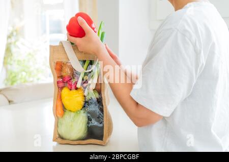 Keine Gesicht Frau hält rote Glocke Papier aus wiederverwendbaren Stoffbeutel Verpackung mit Gemüse auf weißem Küchentisch stehen. Frisches Bio-Gemüse zu Hause Stockfoto
