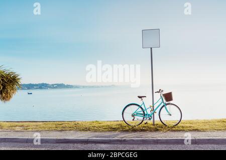 Vintage-Stil helle Fahrrad in der Nähe eines leeren Straßenschild in der Uferpromenade Kai geparkt. Meeresblick Landschaft mit gepflasterten Wanderwegen mit einer Palme bei Sonnen Stockfoto