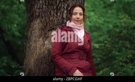 Porträt von jungen hübschen Frau in roter Jacke stehend in der Nähe von Baum im Frühlingswald. Stockfoto