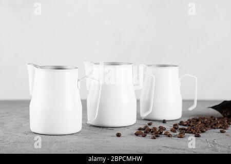 Drei weiße Milchkännchen auf grauem dekorativem Hintergrund. Kaffeebohnen sind in der Nähe verstreut. Stockfoto