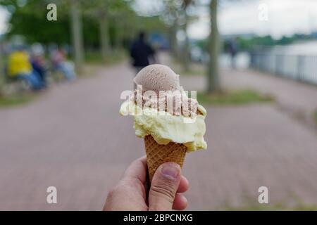 Hand halten 2 Kugeln Schokolade und Vanille Eis auf Waffelkegel mit Unschärfe Hintergrund Park am Flussufer in der Sommersaison. Stockfoto
