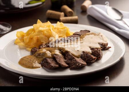 Steak mit Sauce mit Kartoffelchips auf weißem Teller und Weinglas, Kork, Besteck und Serviette Stockfoto