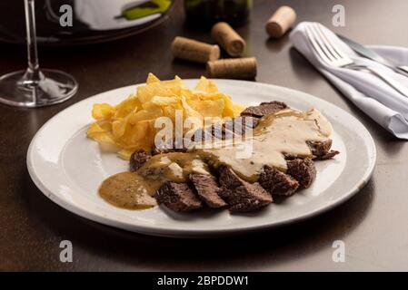 Steak mit Sauce mit Kartoffelchips auf weißem Teller und Weinglas, Kork, Besteck und Serviette Stockfoto