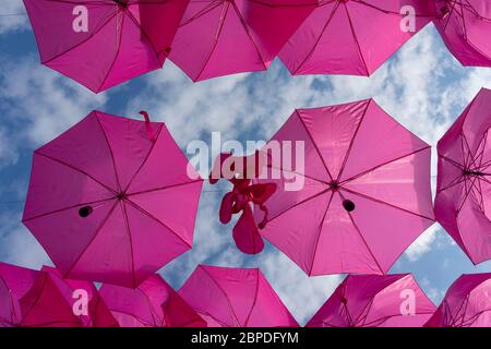 Street Art Installation von farbenfrohen, rosafarbenen Sonnenschirmen, die an einem bewölkten Himmel aufgehängt sind Stockfoto