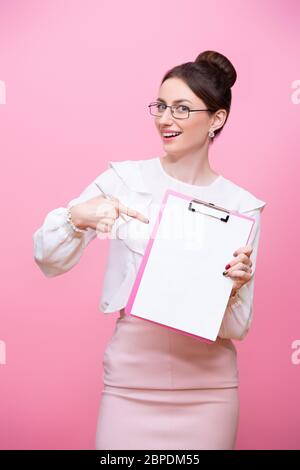 Eine lächelnde junge Frau mit Brille hält einen Ordner in der Hand und zeigt mit einem Finger auf sie. Isoliert auf rosa Hintergrund Stockfoto