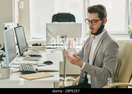Ernst junger schöner Mann mit Maske auf Kinn Spritzen antiseptisch auf die Hände während der Arbeit allein in leerem Büro während Coronavirus Stockfoto