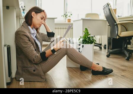 Verärgert junge Frau in stilvollen Casual Anzug sitzen auf dem Boden und lesen Hinweis auf Entlassung in leerem Büro während Coronavirus Quarantäne Stockfoto