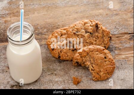 Kleine Glasflasche cremig hofeigene Milch mit einem Strohhalm neben die Hälfte gegessen knusprige Cookies auf einer rustikalen Holzoberfläche, erhöhte Ansicht Stockfoto