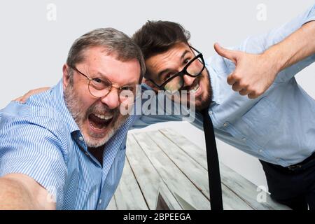 Zwei lächelnde Kollegen machen das Foto zu ihnen selbst im Büro sitzen, glückliche Freunde mit Brille machen Selfie mit Telefonkamera auf weiß Stockfoto