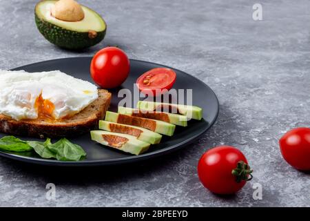 Pochiertes köstliches Ei mit Vollkornbrot, Avocado in Scheiben geschnitten, Kirschtomate und Spinat auf schwarzem Teller auf Steingrund Stockfoto