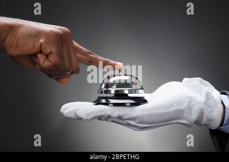 Close-up Kunden Ringing Service Bell Gehalten von Butler Vor grauem Hintergrund Stockfoto
