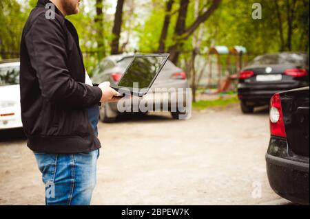 Mann mit einem Laptop auf Parkplatz im Hof in der Nähe von Auto tut Manipulationen mit Cyber-System Konzept Stockfoto