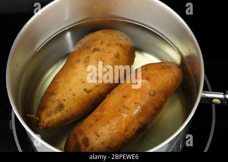 Zwei Orangenkartoffeln, ipomoea batatas, in einer Pfanne mit Wasser zum Kochen bereit. Nahaufnahme von zwei ungekochten, ganzen Süßkartoffeln in Wasser in a de Stockfoto