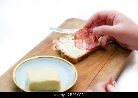 Nahaufnahme der Hand, indem Salami-Wurstscheibe auf Weißbrot mit Butter auf Holzbrett in der Küche Stockfoto