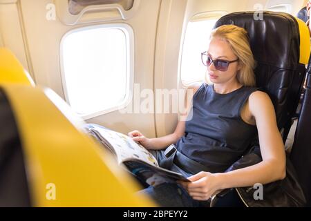 Frau liest Magazin auf dem Flugzeug während des Fluges. Weibliche Reisende Lesung im Passagier Kabine sitzt. Sonne scheint durch Flugzeug Fenster. Stockfoto