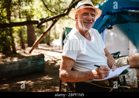 Lächelnder älterer Mann, der vor einem Zelt sitzt und in einem Buch schreibt. Pensionierter Mann, der die Kamera anschaut und lächelt, während er auf dem Campingplatz ein Buch schreibt. Stockfoto