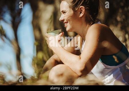 Lächelnde junge Frau, die während des Trekkings Kaffee trinkt. Sportliche Frau, die beim Wandern Ruhe nimmt. Stockfoto