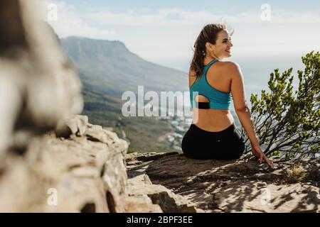 Rückansicht einer gesunden Frau, die auf der Klippe sitzt. Wandererin in Sportbekleidung, die sich nach dem Aufstieg auf dem Gipfel des Berges entspannt. Stockfoto