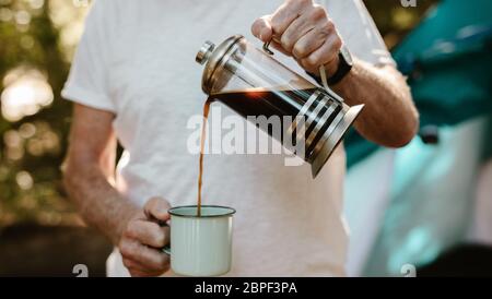 Nahaufnahme eines älteren Mannes, der Kaffee in einer Tasse auf dem Campingplatz gießt. Männlicher Reisender, der Kaffee auf dem Campingplatz getrunken hat. Stockfoto
