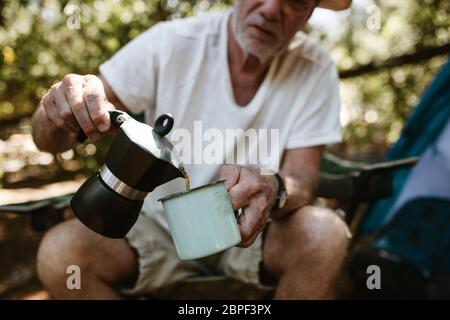 Nahaufnahme eines älteren Mannes, der Kaffee in einer Tasse auf dem Campingplatz gießt. Männlicher Reisender, der Kaffee auf dem Campingausflug getrunken hat. Stockfoto