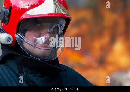 Das Gesicht eines Feuerwehrmundes in einem Helm auf einem Hintergrund des Feuers. Ein Feuer löschen. Stockfoto