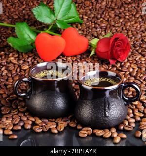 Zwei Tassen schwarzen Kaffee mit Schaum auf dem Hintergrund von den gebratenen Kaffeebohnen, kleine Herzen und eine rote Rose. Für Karten zum Valentinstag. Stockfoto