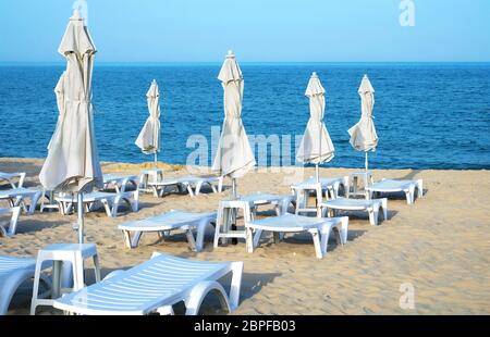 Leere Sonnenliegen und geschlossene Sonnenschirme an einem einsamen Sandstrand am klaren Tag. Stockfoto