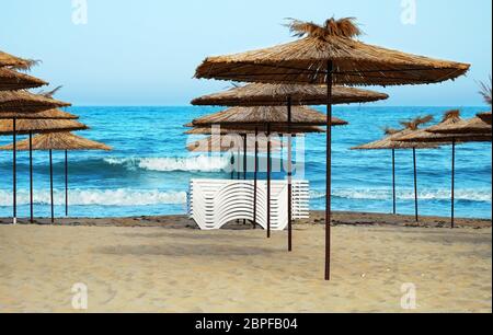 Leerer Strand im Badeort mit gefalteten Sonnenliegen und Holzschuppen mit Strohdach auf einem Hintergrund des blauen Meeres. Stockfoto