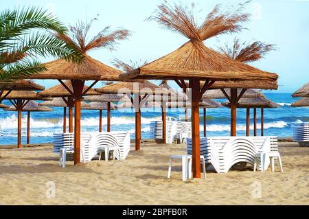 Leerer Strand im Badeort mit gefalteten Sonnenliegen und Holzschuppen mit Strohdach auf einem Hintergrund mit blauen Meereswellen. Stockfoto