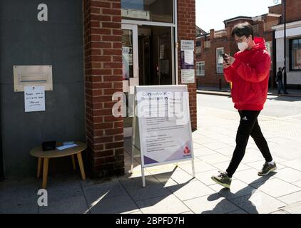 Ein junger Koreaner, der eine Maske trägt und auf sein Mobiltelefon schaut, während er an einer NatWest-Bank in der New Malden High Street vorbeigeht. Stockfoto