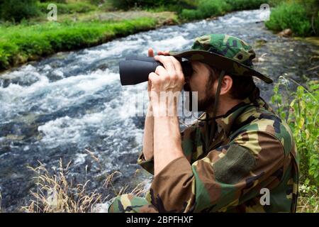 Der Naturforscher in Militäruniform mit Fernglas ist auf Vogelbeobachtungsmission in der Natur am Flussufer. Stockfoto