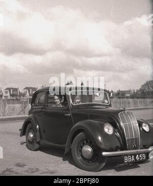 Draußen auf einem leeren Parkplatz, vielleicht an der Küste, sitzt ein erwachsenes Paar in einem beliebten Auto der Zeit, einem Ford Anglia, mit dem Mann auf dem Fahrersitz, England, Großbritannien, 1950er. Stockfoto