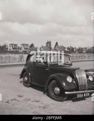 Draußen auf einem leeren Schotterparkplatz, vielleicht an der Küste, sitzt ein erwachsenes Paar in einem beliebten Auto der Zeit, einem Ford Anglia, mit dem Mann auf dem Fahrersitz, England, Großbritannien, 1950er. Stockfoto