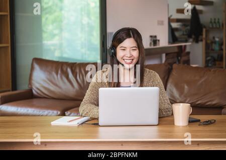 Asiatische Geschäftsfrau, die mit einem technischen Laptop und einem Kopfhörer arbeitet, um von zu Hause aus per Videokonferenzanruf, Startups und Geschäftsinhaber im Haus zu arbeiten Stockfoto