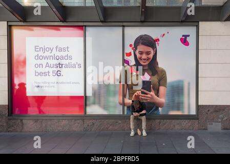 Ein junger Mann sitzt Kopf unten und schaut auf sein Telefon direkt unter einem Telstra 5G Poster, während eine lächelnde Frau auf ihr Telefon schaut. Stockfoto