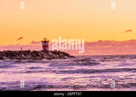 Rot-weiß gestreifter Leuchtturm bei einem lebhaften Sonnenuntergang. Wasser trifft die schwarzen Felsbrocken in Wellen. Dramatischer Himmel in goldener Farbe. Scheveningen, die Neth Stockfoto