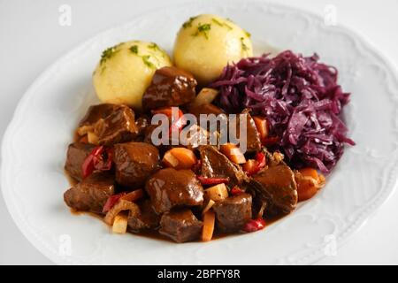 Würziger Wildschwein oder Reh Gulasch in einem reichhaltigen Soße mit Klößen und Rotkohl auf einem weißen Teller serviert Stockfoto