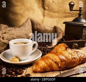 Frisch gebackene Croissant mit einer Tasse Kaffee und Macaron vor dem Hintergrund von gerösteten Kaffeebohnen in einem sackleinen Sack mit Vintage Kaffeemühle Stockfoto