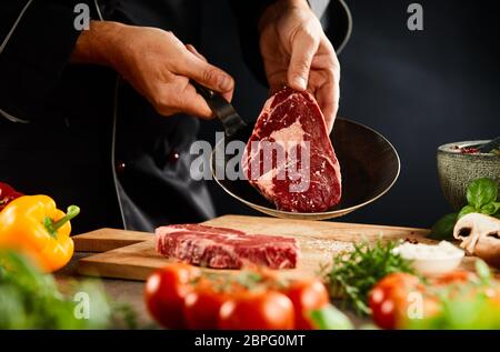Koch, ein rohes Steak in einer Pfanne für Schmoren oder Grillen mit frischem Gemüse im Vordergrund. Stockfoto