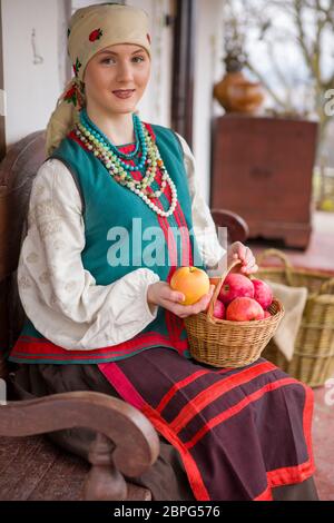 Ein schönes Mädchen in Nationalkleid sitzt. Mit Äpfeln in einem Korb. In alten Kleidern des 19th. Jahrhunderts. Mit Kranz und Bändern. Gegen die Stockfoto