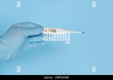 Hand in Handschuhe hält Digitalthermometer vor blauem Hintergrund. Gesundheitskonzept und medizinisches Konzept. Stockfoto