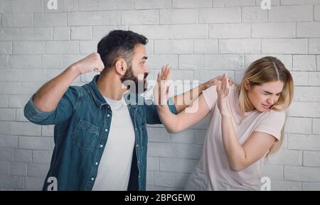 Wütender Mann, der Faust schwingt und Frau hält, verängstigte Frau versteckt sich vor Schlägen Stockfoto