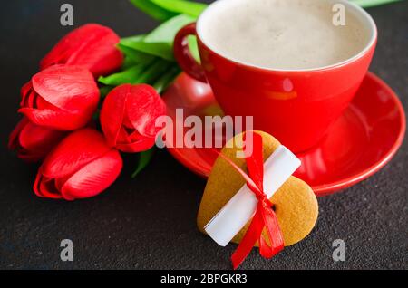 Romantische Komposition für Valentinstag, Geburtstag oder Muttertag. Rote Kaffeetasse, Cookie in der Form eines Herzens mit einem Hinweis und roten Tulpen. Stockfoto